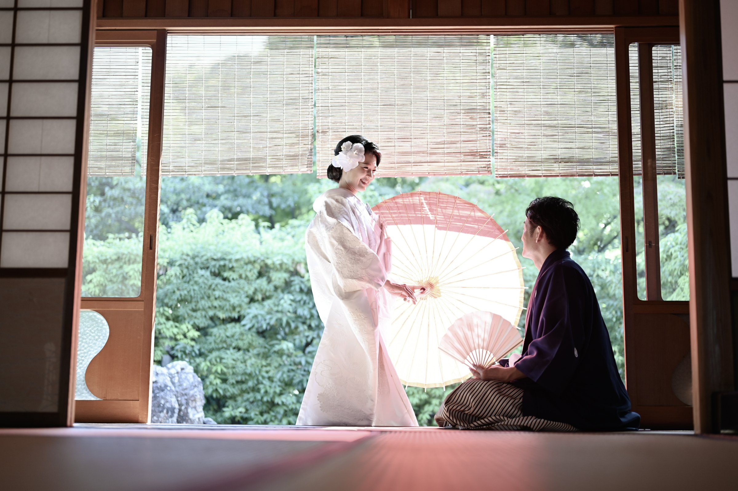 障子の間から庭園を望む縁側で、白い和傘を差す白無垢の新婦と紫の羽織袴の新郎