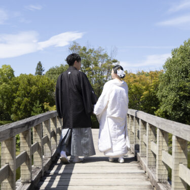 青空の下、木の橋を並んで歩く紋付袴の新郎と白無垢の花嫁の後ろ姿。白鳥庭園で撮る和装ロケーションフォト