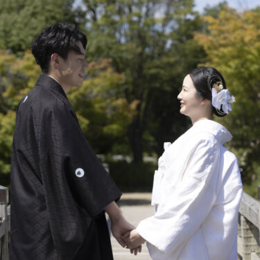 橋の上で手をつなぎ見つめ合う、紋付の新郎と胡蝶蘭の髪飾りを付けた白無垢の花嫁。白鳥庭園の和装前撮り