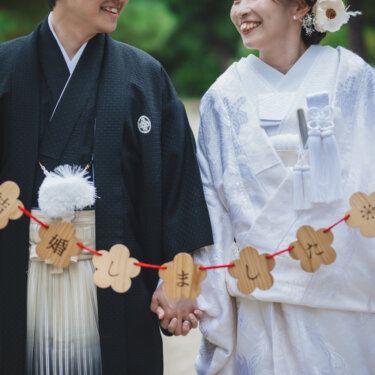 '結婚しました'の文字を梅形の木札でつないだ赤い糸のガーランドを持ち、手をつなぐ紋付袴の新郎と白無垢の花嫁。結婚報告にも使える和装の結婚写真