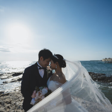 風になびくロングベールに包まれ、岩場で額を寄せ合う新郎新婦。知多半島の青い海で撮るロケーションフォト