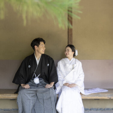 松の枝越しに、茶屋の軒下のベンチに並んで座る紋付袴の新郎と白無垢の花嫁。日本庭園で撮る和装の結婚写真