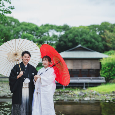 池のほとりで白と赤の和傘を差して並ぶ紋付袴の新郎と白無垢の花嫁。和風の建物を背景にした白鳥庭園の和装前撮り