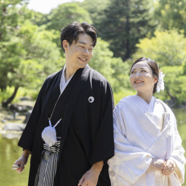 池と松を背に並んで歩き、笑い合う紋付袴の新郎と白無垢の花嫁。白鳥庭園の夏の緑に包まれた和装フォトウェディング