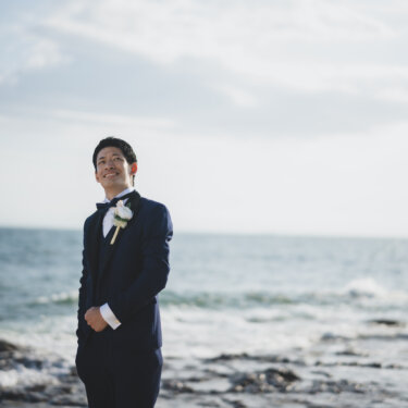海を背にブートニアを付けて笑う、ネイビーのタキシードの新郎のソロカット。知多半島の海辺で撮る洋装前撮り