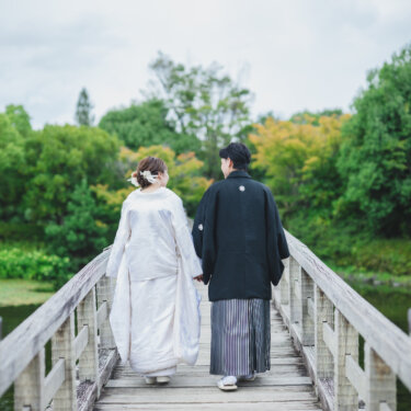 池に架かる木の橋を手をつないで渡る、白無垢の花嫁と紋付袴の新郎の後ろ姿。白鳥庭園で撮る和装の結婚写真