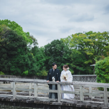 曇り空の下、池に架かる木の橋の欄干にもたれて並ぶ紋付袴の新郎と白無垢の花嫁。緑に囲まれた日本庭園での和装前撮り