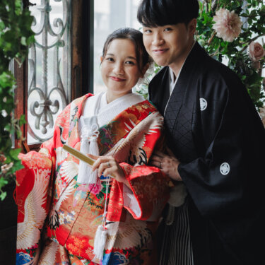 アイアン飾りの窓の前で扇子を手に、肩を寄せて笑う赤い色打掛の花嫁と紋付袴の新郎。アトリエスタジオで撮る和装前撮りの縦構図