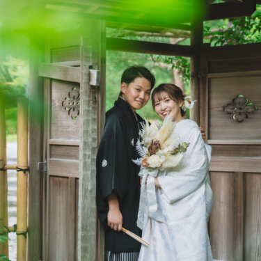 竹垣と木の門の前で寄り添い、ブーケを手に微笑む白無垢の花嫁と紋付袴の新郎。白鳥庭園の風情ある門で撮る和装前撮り