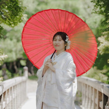 赤い和傘を差して木の橋に立ち、やわらかく微笑む白無垢の花嫁のソロカット。白鳥庭園での和装前撮り