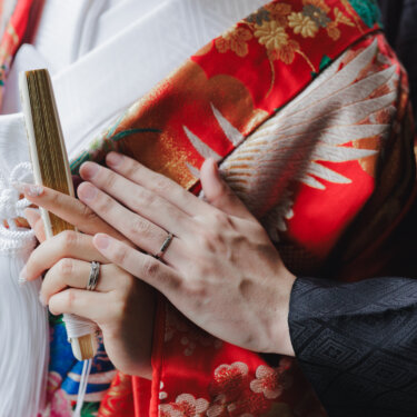 赤い色打掛の胸元で、扇子を持つ花嫁の手に新郎がそっと手を重ね、結婚指輪が光る手元のカット。和装前撮りで残したい定番ショット