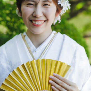 金色の扇子を手に満面の笑みを見せる、白無垢に白い髪飾りを合わせた花嫁のアップの前撮り写真