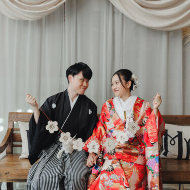 結婚しましたのガーランドを手に、ベンチで顔を見合わせて笑う紋付袴の新郎と色打掛の花嫁。アイテムを使った和装のスタジオフォト