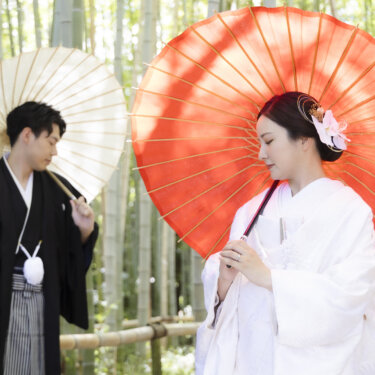 竹林を背に、白い和傘の新郎と赤い和傘の白無垢の花嫁が並ぶ構図。白鳥庭園の竹垣の前で撮る和装前撮り