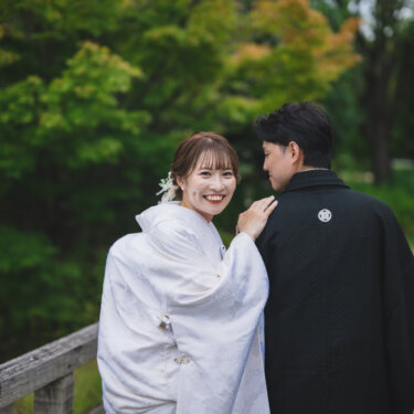 木の橋の上で新郎の背中に手を添え、振り返って笑う白無垢の花嫁。白鳥庭園の緑を背景にした和装前撮りのポーズ