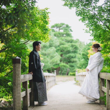 緑のもみじに囲まれた木の橋で、欄干にもたれて向き合う紋付袴の新郎と白無垢の花嫁。日本庭園で撮る和装の結婚写真