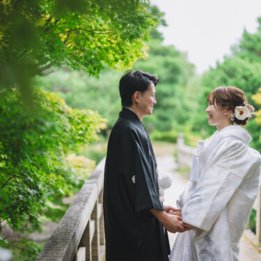 青もみじの枝越しに、橋の上で手を取り見つめ合う紋付袴の新郎と白無垢の花嫁。白鳥庭園の緑が美しい和装前撮り