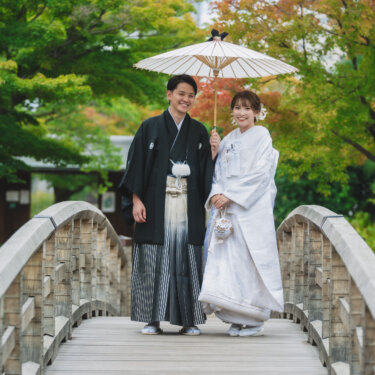 色づき始めたもみじを背に、太鼓橋の上で白い和傘を差して並ぶ紋付袴の新郎と白無垢の花嫁。白鳥庭園の和装前撮り
