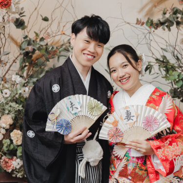 夫と妻の文字入り扇子を手に、ドライフラワーの装花の前で笑顔を見せる紋付袴の新郎と赤い色打掛の花嫁。名古屋のスタジオで撮る和装前撮り