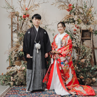 花と枝のアレンジに囲まれたキリムラグの上に並んで立つ、紋付袴の新郎と裾を広げた赤い色打掛の花嫁。アトリエスタジオの和装フォトウェディング