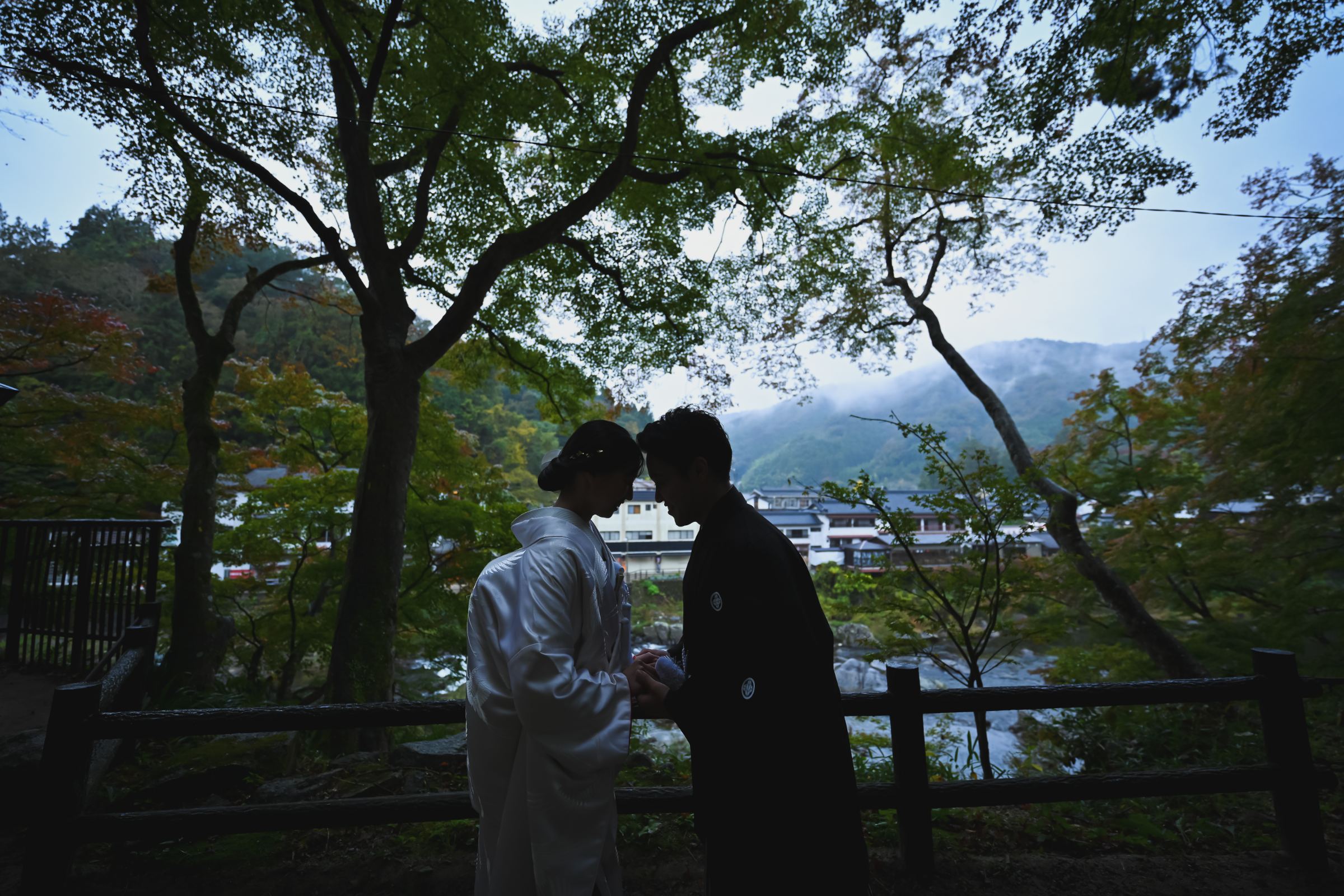山と川霧を望む夕暮れ、額を寄せ合うシルエットの新郎新婦、白無垢と黒紋付の香嵐渓の里山風景。