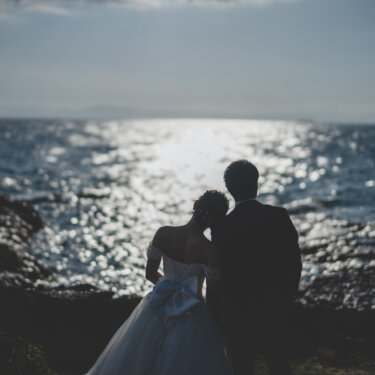 きらめく海を眺め、寄り添って立つ新郎新婦の後ろ姿のシルエット。夕方の知多半島の海で撮る前撮り