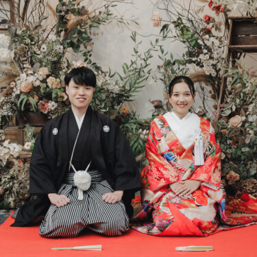 赤い毛氈の上に正座して並び、穏やかにほほえむ紋付袴の新郎と色打掛の花嫁。ドライフラワーの装花が映えるスタジオの和装結婚写真