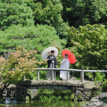 池にかかる石橋の上で白と赤の和傘を差して立つ新郎新婦を、白鳥庭園の緑とともに遠くから写した前撮り
