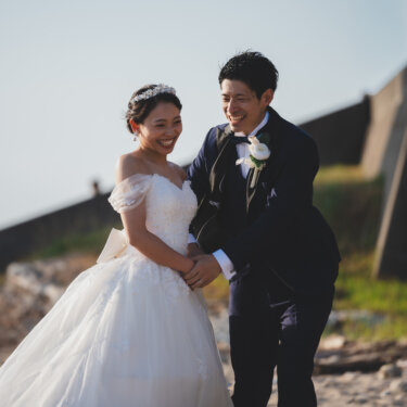 海辺の堤防の前で、花嫁の手を取って楽しそうに笑う新郎。オフショルダードレスで撮る洋装前撮り
