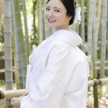 竹林の前で振り返って微笑む、白い花の髪飾りを付けた白無垢の花嫁のソロカット。和装ロケーションフォトの一枚