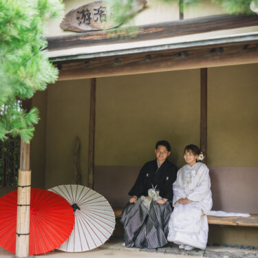 赤と白の和傘を立てかけた東屋のベンチに並んで座る、紋付袴の新郎と白無垢の花嫁。白鳥庭園で撮るほっこりした和装前撮り