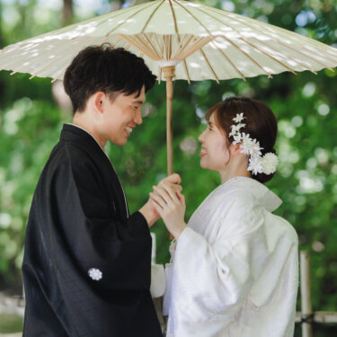 白い和傘の下で手を重ね、見つめ合って微笑む黒紋付袴の新郎と白無垢の花嫁の結婚写真