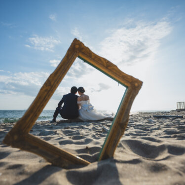 砂浜に立てたゴールドの額縁の中に、灯台のそばで並んで座る新郎新婦を収めた構図。野間埼灯台のビーチで撮る前撮り