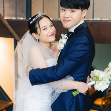 階段の下でぎゅっとハグし、カメラに笑顔を向けるベール姿のウェディングドレスの花嫁と紺のタキシードの新郎。名古屋のスタジオフォトウェディング