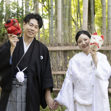 竹垣の前で手をつなぎ、天狗と狐のお面を手に笑う新郎新婦。白鳥庭園で撮る遊び心のある和装前撮りの小物使い