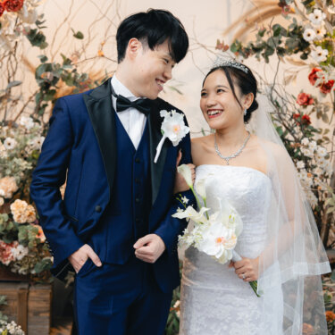 ドライフラワーの装花を背に、カラーのブーケを持つマーメイドドレスの花嫁と紺のタキシードの新郎が笑い合う、スタジオ前撮りの一枚