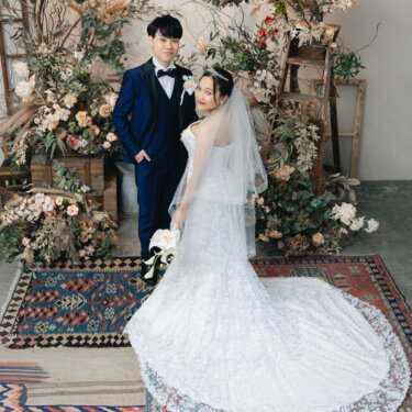 キリムラグに長いレースのトレーンを広げたウェディングドレスの花嫁と、後ろに立つ紺のタキシードの新郎。アトリエスタジオで撮る洋装前撮り