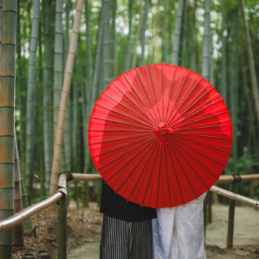 竹林の小道で赤い和傘に顔を隠し、足元だけをのぞかせる新郎新婦。和傘を使った遊び心のある和装前撮りのポーズ