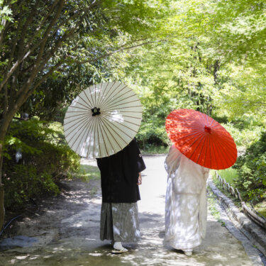 緑のトンネルの小道を、白と赤の和傘をさして歩く新郎新婦の後ろ姿。夏の日本庭園で撮る和装フォトウェディング