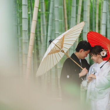竹林で白と赤の和傘を差し、おでこを寄せて笑う紋付袴の新郎と白無垢の花嫁。緑の竹に囲まれた和装前撮り