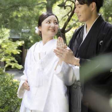 松の木の下で、指輪を付けた手を見せて新郎に微笑む白無垢の花嫁。白鳥庭園の緑の中で撮る和装前撮り