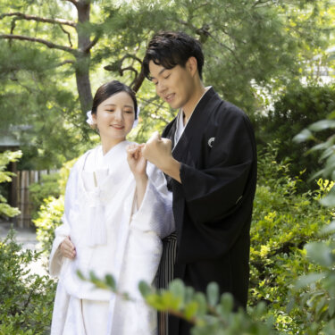 庭木の間で寄り添い、手を合わせて笑い合う紋付袴の新郎と白無垢の花嫁。自然体で撮る和装の結婚写真