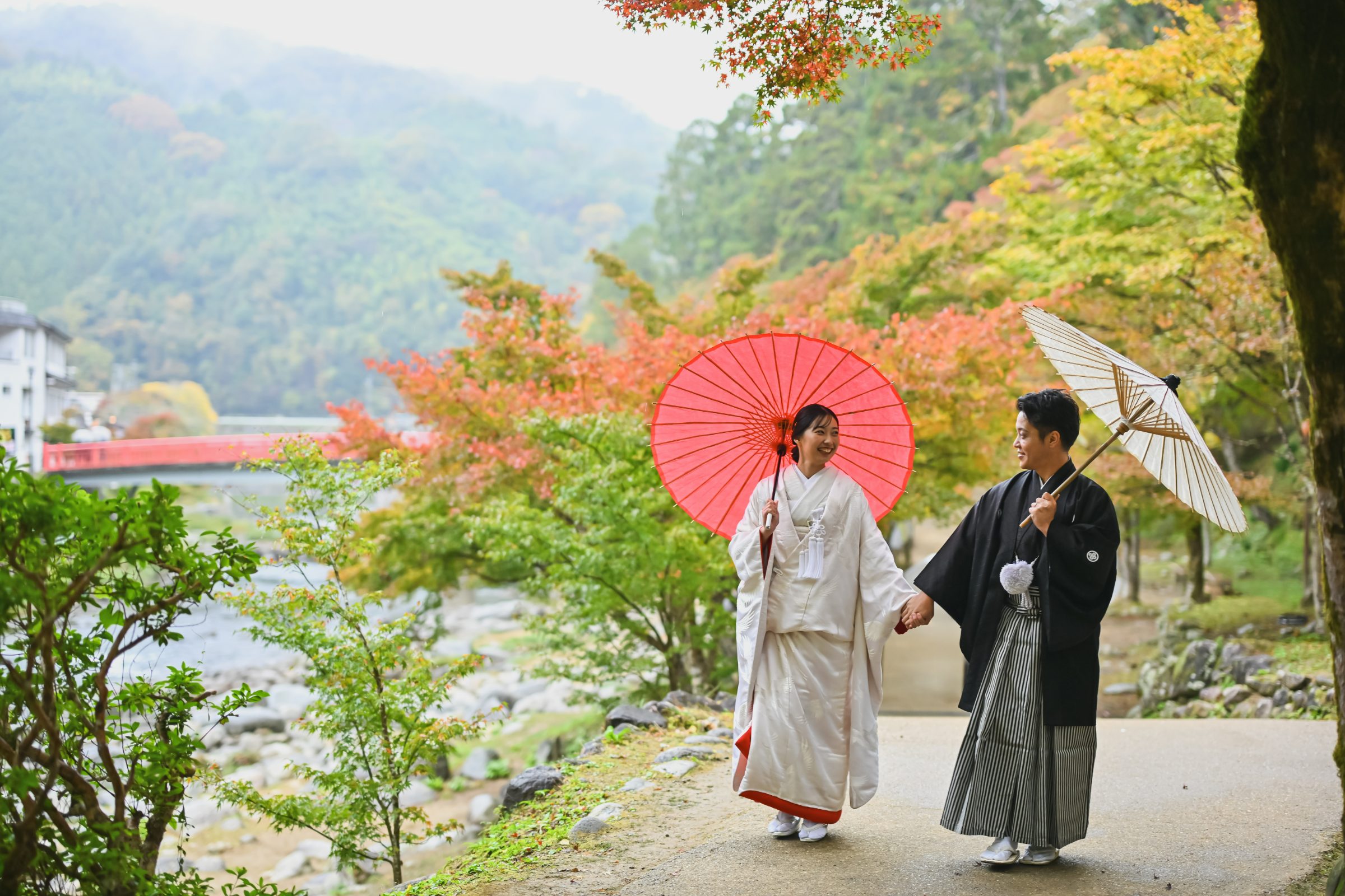 朱色と白の和傘をさし、手をつないで歩く白無垢姿の花嫁と黒紋付袴姿の新郎。紅葉の山と赤い欄干の橋を望む渓流沿いの一枚。