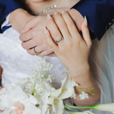 花嫁の胸元で新郎の手に重ねた結婚指輪の手元と、胡蝶蘭とカラーの白いブーケ。洋装のフォトウェディングで残したいクローズアップ