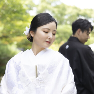 目を伏せて微笑む白無垢の花嫁と、奥でうつむく紋付の新郎。白鳥庭園の緑をぼかして撮る和装フォトウェディング