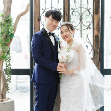 アイアン飾りの窓辺とグリーンの前で、白いブーケを手に寄り添うウェディングドレスの花嫁と紺のタキシードの新郎。名古屋のスタジオ前撮り