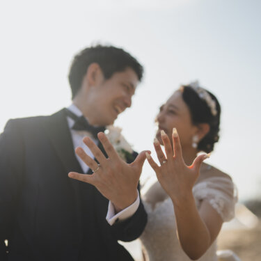 灯台を背に、結婚指輪を付けた手を開いて見せ合う新郎新婦。笑顔があふれる野間埼灯台のウェディングフォト