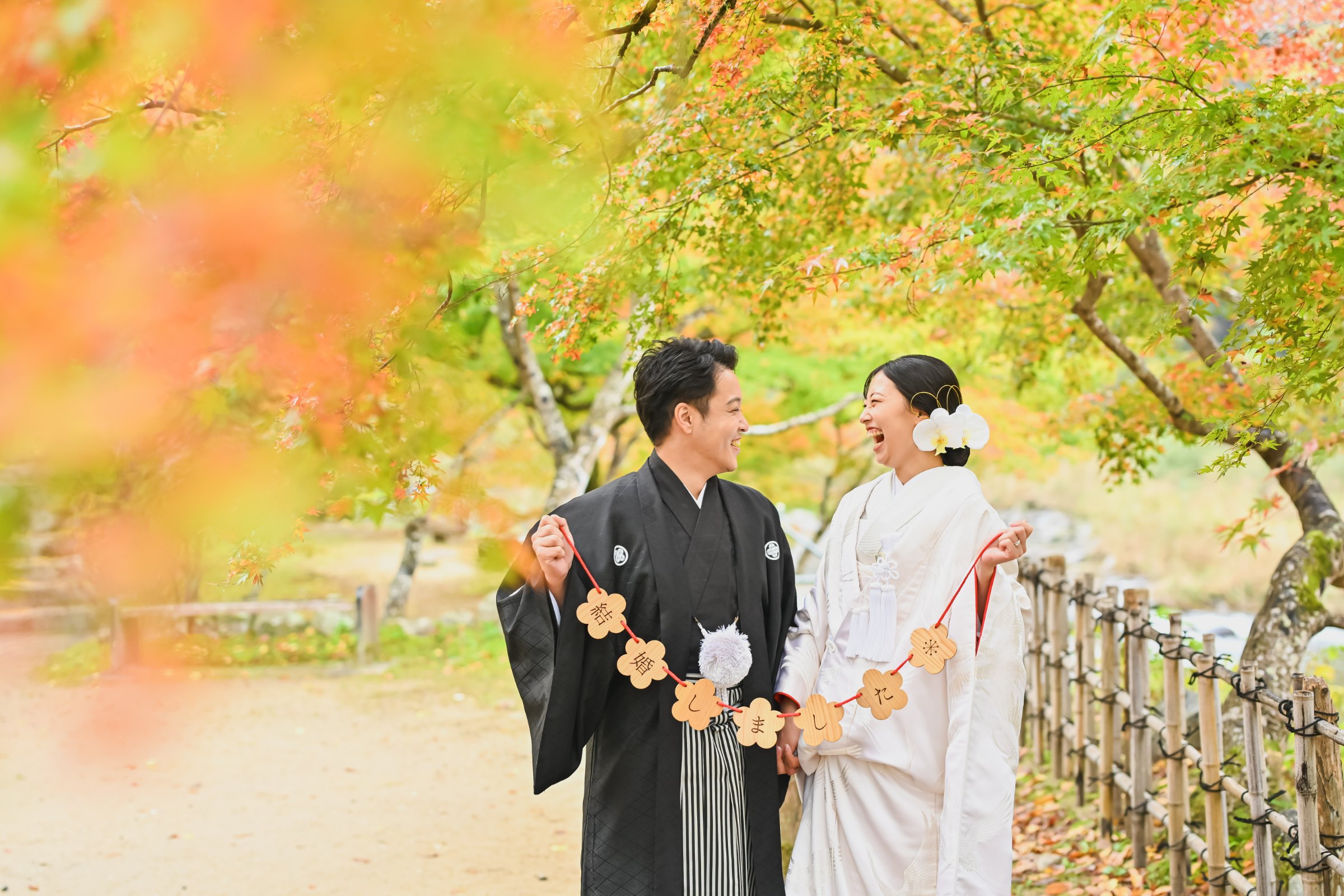 花型のガーランドを掲げて大笑いする黒紋付袴姿の新郎と白無垢姿の花嫁。紅葉の並木道を背景にした賑やかな一枚。