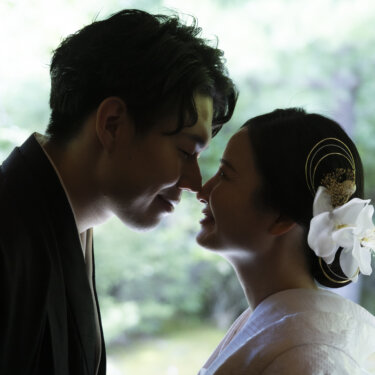 額を寄せ合い目を閉じる紋付の新郎と白無垢の花嫁の横顔アップ。日本庭園の緑を背景にした和装の結婚写真