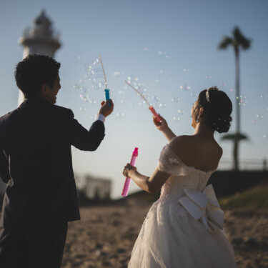 灯台とヤシの木を背景に、シャボン玉を飛ばして遊ぶ新郎新婦の後ろ姿。夕方の海辺で撮るかわいい前撮り